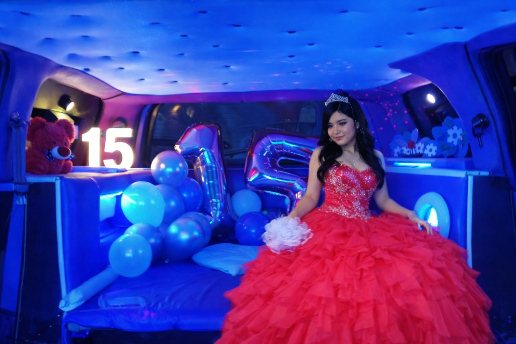 Interior de Hummer rosa decorada para fiesta de 15 años con iluminación LED azul, globos y número 15 iluminado, quinceañera con vestido rojo sentada en el sofá.