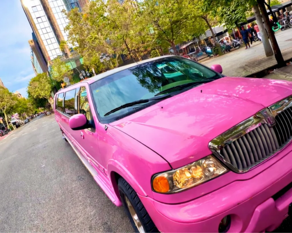 Una limusina estilo hummer Rosa circula por la carretera, con modernos y altos edificios de Barcelona al fondo, mostrando un paisaje urbano elegante y vibrante